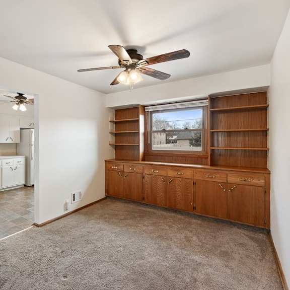 an empty living room with a ceiling fan and a kitchen in the background
