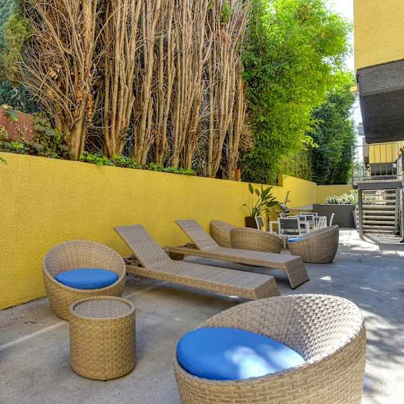 community lounge area with wicker seats with blue cushions, wicker lounge chairs, trees, and yellow wall at croft plaza apartments, west hollywood