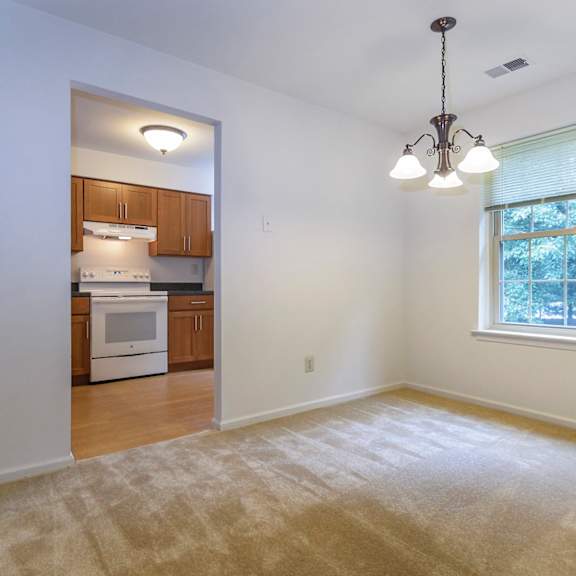 a dining room with carpet flooring, an overhead chandelier, and a window for natural lighting with an entry to a kitchen.