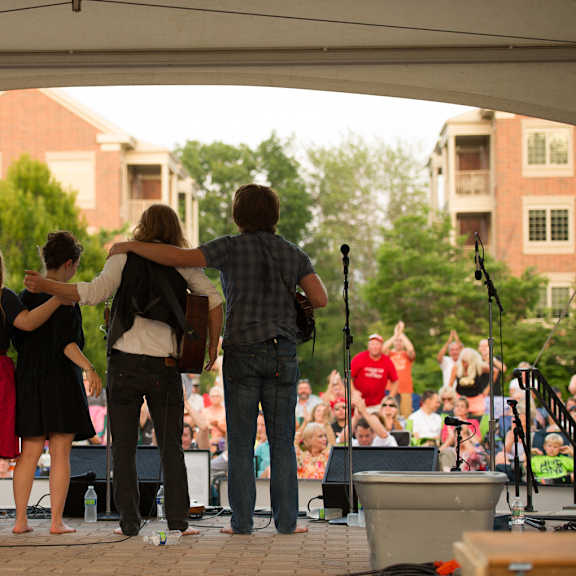 a concert at eagleview town center showing the backside of four performers with their instruments on an outdoor covered stage with a crowd of people watching them.