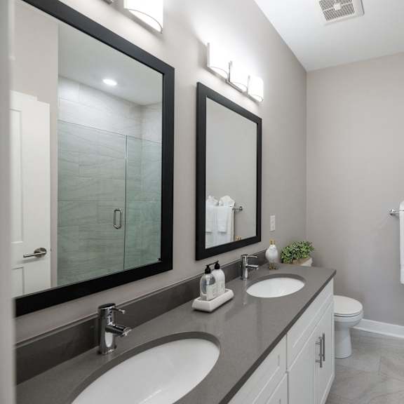 a bathroom with double vanity sinks, two large mirrors, a toilet, and a swinging glass door to a stand up shower with tile walls.