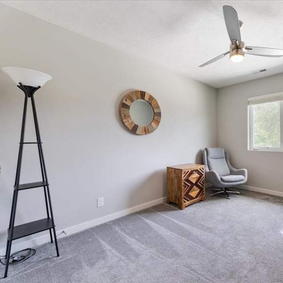 a room with a grey carpet, a ceiling fan, a window, and a wooden box on the floor.