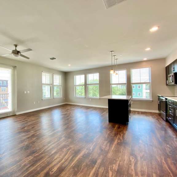 a large open living room with six windows, double doors to a balcony, a ceiling fan, and hardwood-style flooring near a kitchen with an island bar.