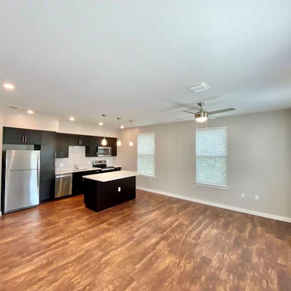 a large and open living space with 3 windows, hardwood-style flooring, and a ceiling fan leading a kitchen with stainless steel appliances.
