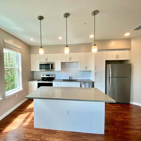 a well-lit open concept kitchen with two large windows, recessed and pendant lighting, hardwood-style flooring, white cabinets, subway tile backsplash, and stainless steel appliances.