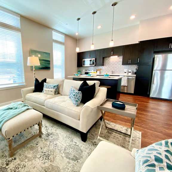 a living room with hardwood style flooring with two windows with open access to a kitchen with an island bar and hanging pendant lights.