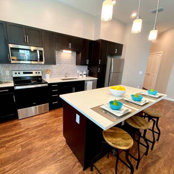 a large kitchen with a breakfast bar, three pendant lights, stainless steel appliances, hardwood-style flooring, a window, and black cabinets.