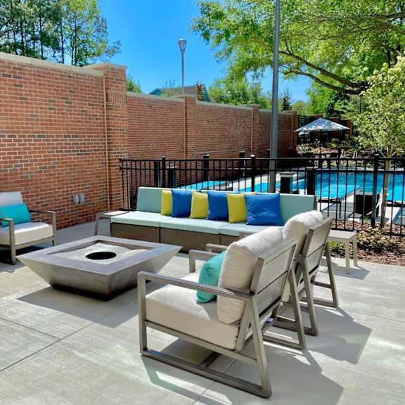 an outdoor community lounge with 4 comfortable chairs, a sofa, and a fire pit near a gated swimming pool.