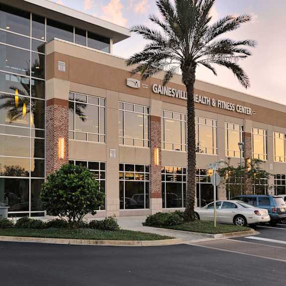 an exterior view of the gainesville health & fitness center with palm trees and a parking lot near the flats at tioga town center apartments.