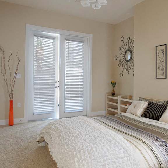 a staged bedroom with plush carpet flooring, ample natural light, and double doors to a private outdoor balcony.