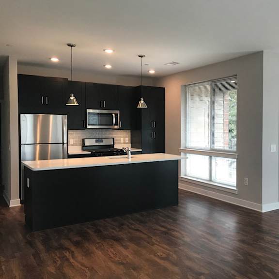 a kitchen with black cabinets, stainless steel appliances, pendant and recessed lighting, a window, and hardwood-style flooring with open access to the living room  at keva flats, exton, 19341.