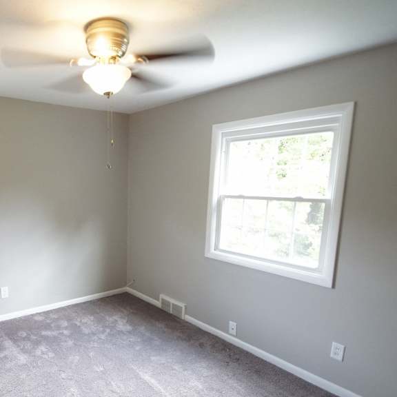 a room with a ceiling fan and two windows and grey carpet.