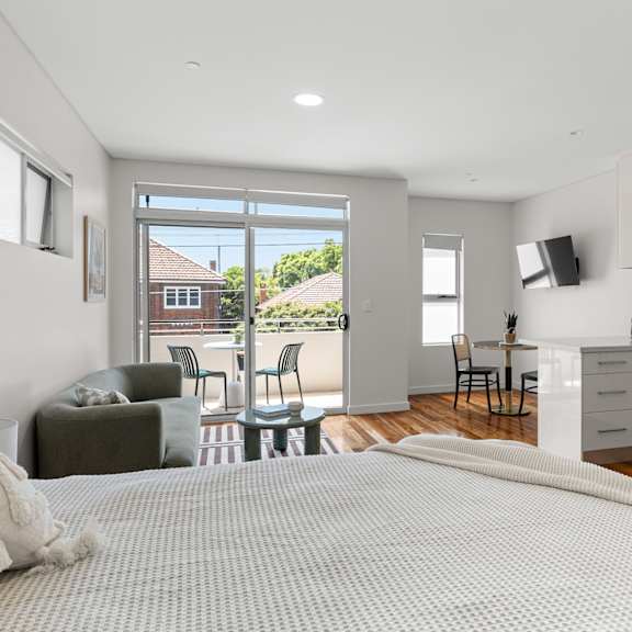 a bedroom with a large bed and a kitchen area in the background.