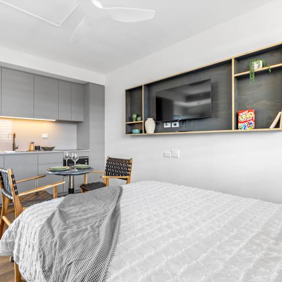 a modern bedroom with a bed, chair, and a kitchen area in the background.