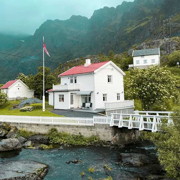 Thumbnail 3 of 3 A white house with a red roof is situated by a body of water with a bridge leading to it.