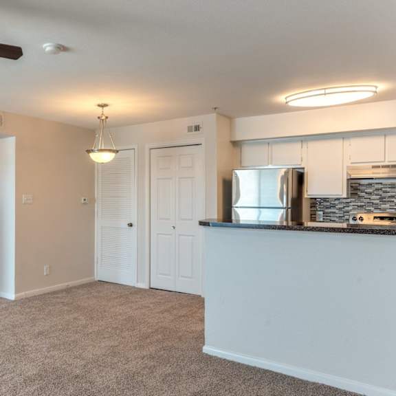 a living room with a ceiling fan and a kitchenette in the background.