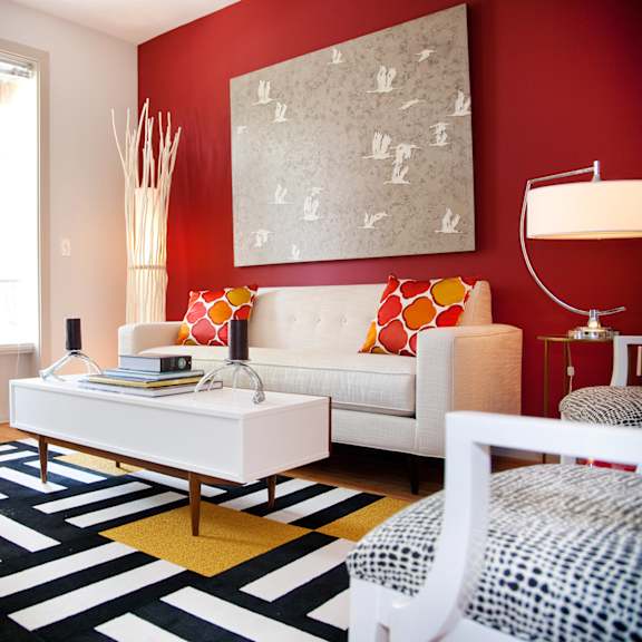 a white sofa with red and orange pillows sits in a room with a red wall and a white chair.