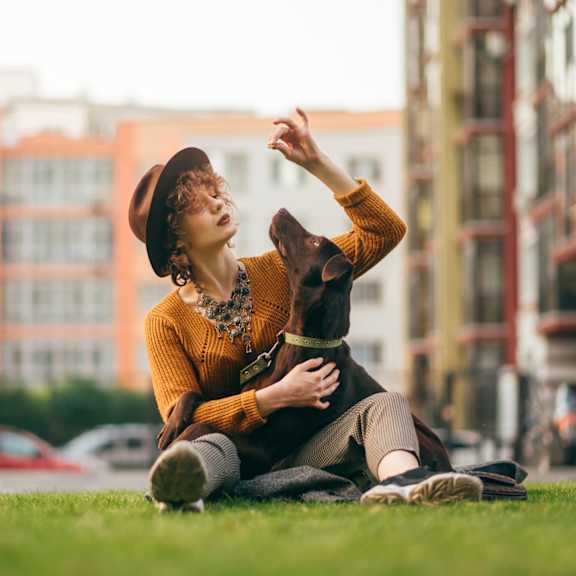 a woman in a yellow sweater and brown hat is sitting on the grass in front of a row of buildings.