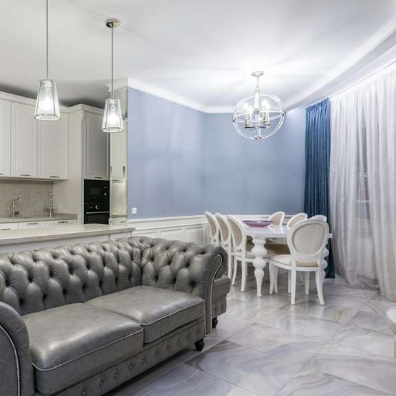 a grey tufted sofa sits in a kitchen with white cabinets and a marble floor.