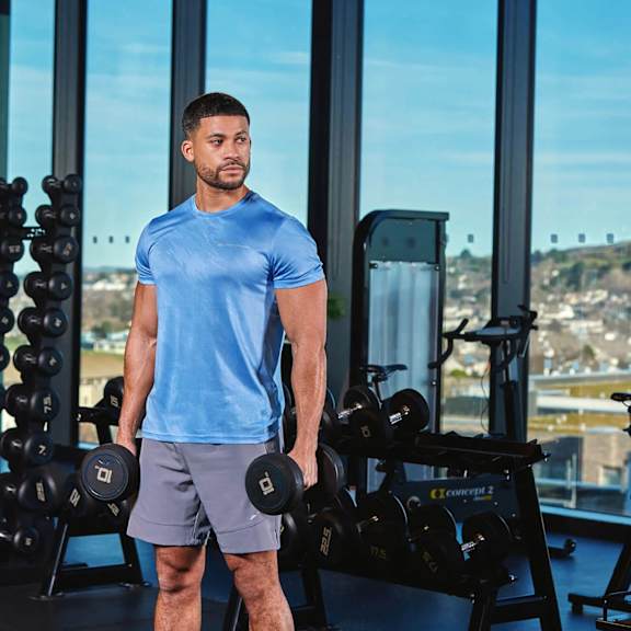 a man in a blue shirt and grey shorts is standing in a gym holding a dumbbell.