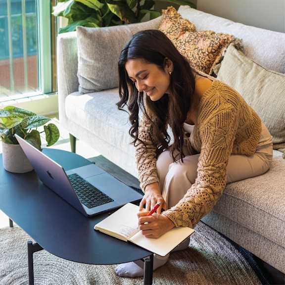 A woman is sitting on a couch with a laptop and a notebook.