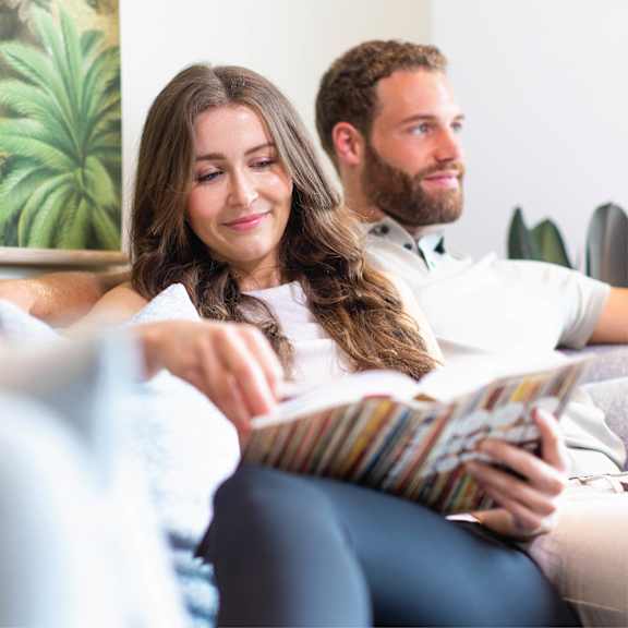 A woman is reading a book while sitting on a couch with a man beside her.