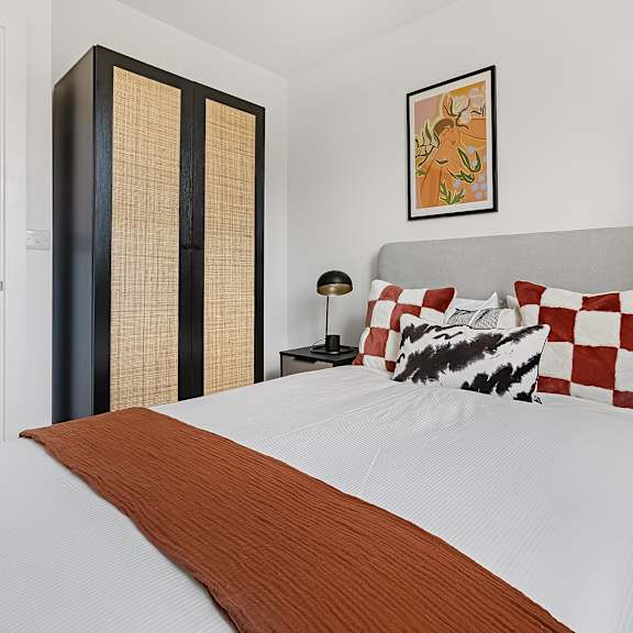 Thumbnail 1 of 5 A bed with a white comforter and a brown throw sits in a room with a tall wardrobe and a lamp.