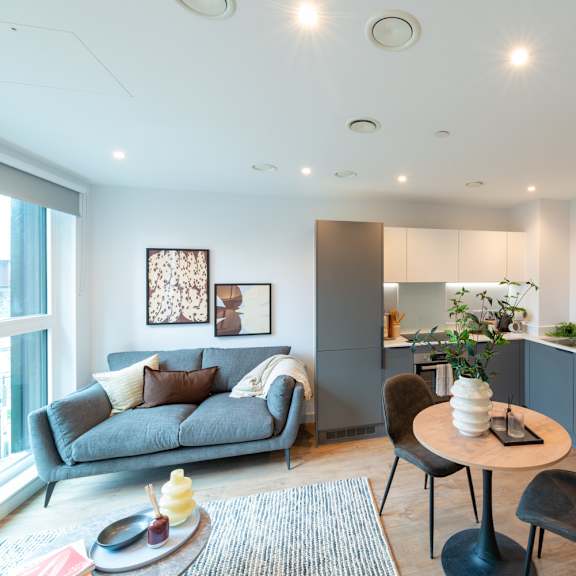 Thumbnail 3 of 11 A modern living room with a grey couch, a round table with a vase of flowers, and a kitchen area in the background.