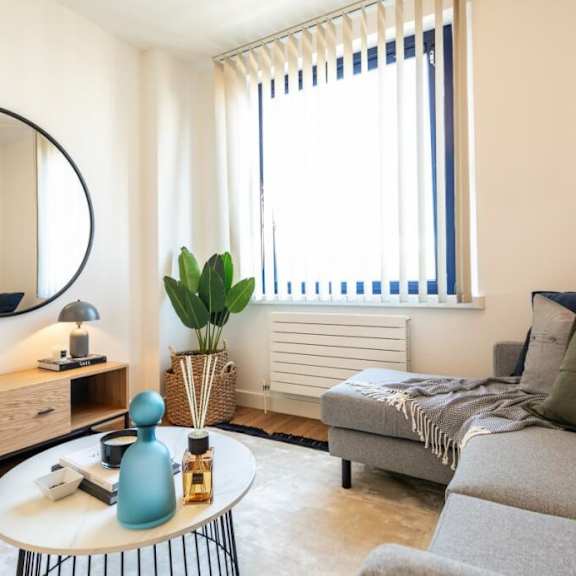 Thumbnail 1 of 3 A living room with a grey couch, a round mirror, a wooden cabinet, a table with a blue vase, and a window with blinds at The Keel Apartments, Liverpool