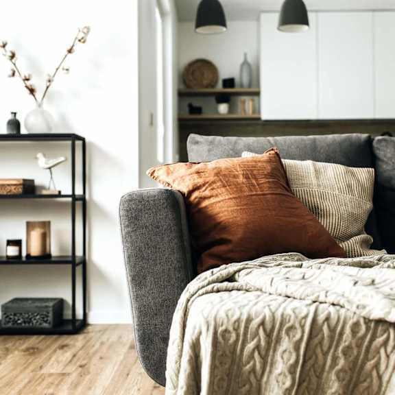 Thumbnail 2 of 3 A grey couch with a blanket and pillows in a room with a shelf and decorations.