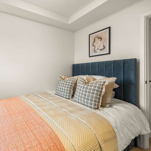 Thumbnail 4 of 5 A bed with a white comforter and orange throw is in a room with a lamp and picture on the wall.