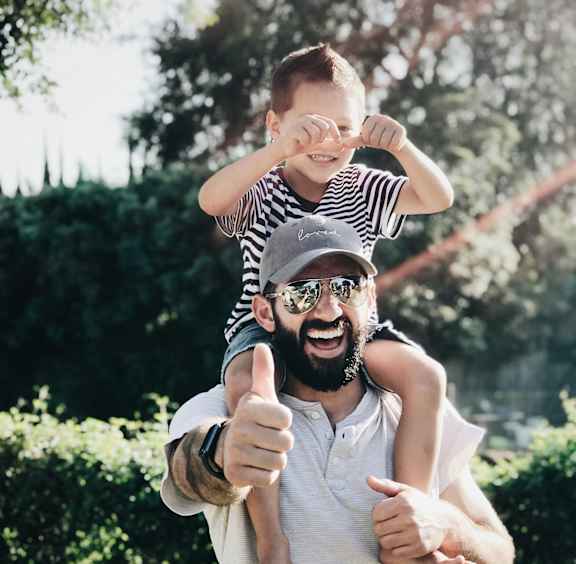 A man is giving a thumbs up while holding a child on his shoulders.