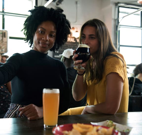 two women sitting at a table drinking beer and looking at a cell phone