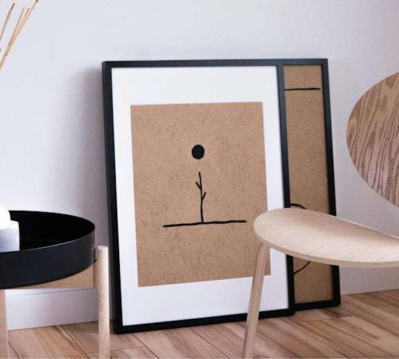 A framed artwork with a brown background and a black border is on a wall next to a round wooden table with a candle and a vase with reeds.