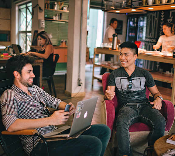 Two men are sitting in a cafe, one is using a laptop and the other is holding a phone.