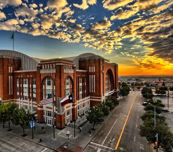 A large red brick building with a curved roof and a flag on top is surrounded by trees and a street in front.