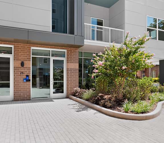 A street view of a building with a brick facade and a sidewalk lined with plants and a lamp post.