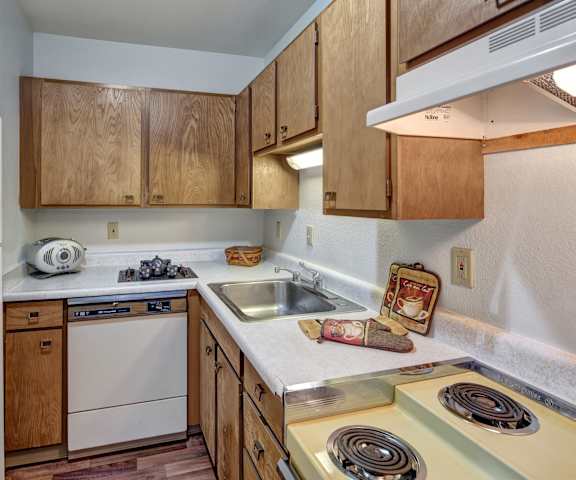 A cozy kitchen with white appliances, kitchen utensils, overhead lighting, light countertops with lots of room, and large wooden cabinets at the Alaskan Apartment Homes. 