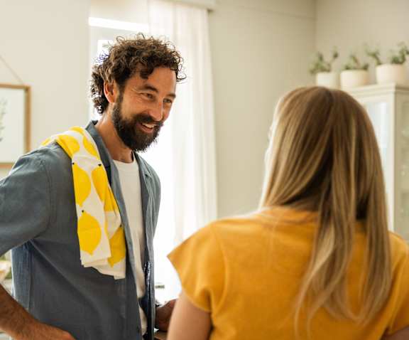 A man with curly hair and a beard is standing in front of a woman, both are smiling.