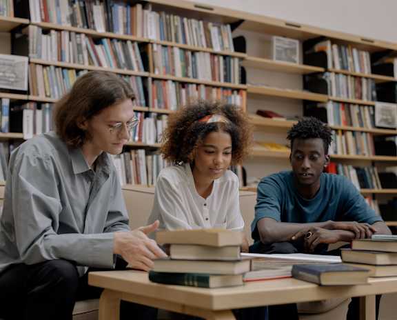 Three people are studying in a library with bookshelves in the background.