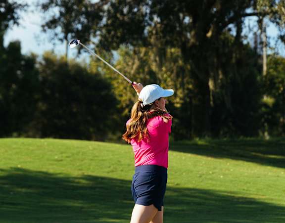 A woman in a pink shirt and blue shorts is playing golf on a sunny day.