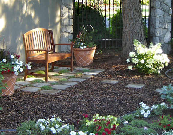 A wooden chair sits in a garden with a stone pathway leading to it.