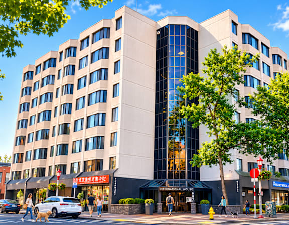 A large building with a curved glass facade is surrounded by trees and people walking on the sidewalk.