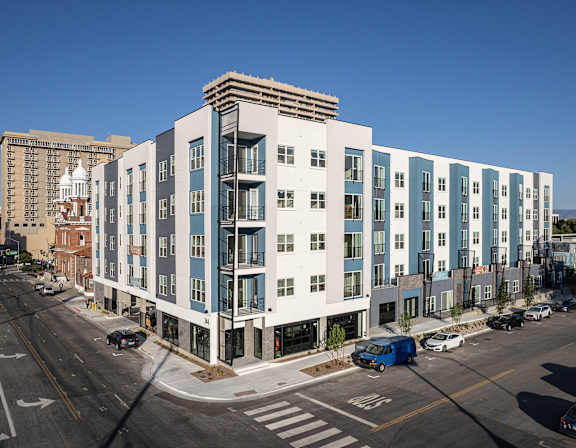A modern multi-story building with a blue and white exterior is situated on a street corner with cars and a traffic light visible.