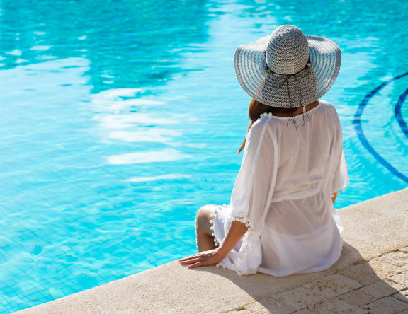 a woman in a white dress and a straw hat sits on the edge of a pool