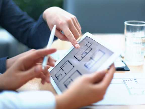 A person is using a tablet to view a floor plan on a table with another person pointing at it.