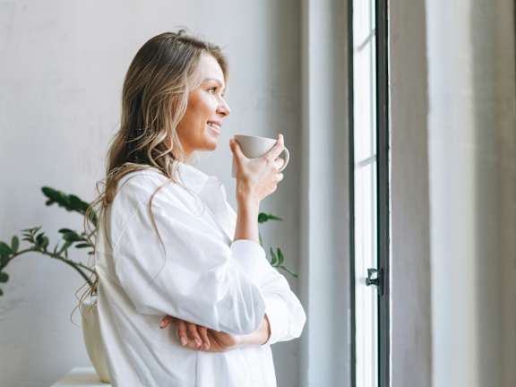 A woman in a white shirt is holding a cup and standing by a window.
