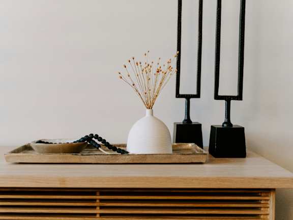 A white vase with dried flowers sits on a table next to two black objects.