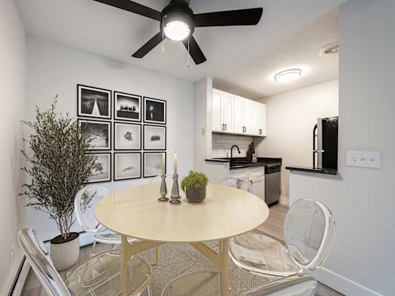 A dining room with a white table and chairs, a ceiling fan, and a kitchen area in the background.
