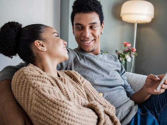 A couple is sitting on a couch, with the woman wrapped in a beige knitted blanket and the man holding a smartphone.